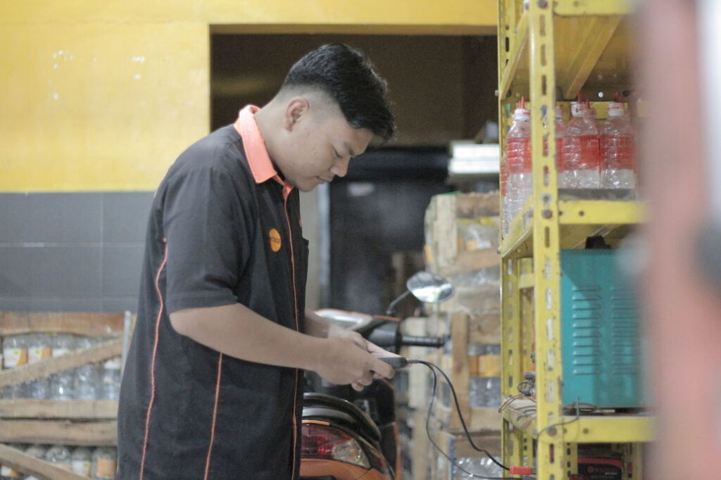 Teknisi toko aki sedang memeriksa perangkat dengan latar belakang rak berisi botol dan peralatan bengkel, menunjukkan suasana kerja di toko aki.